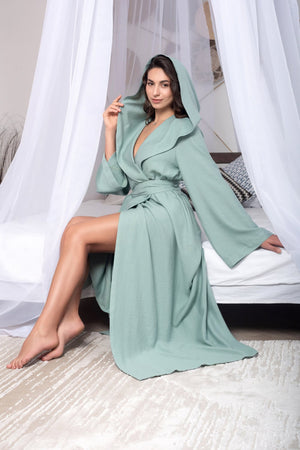 Woman wearing a long light green robe sitting on a bed with white curtains.