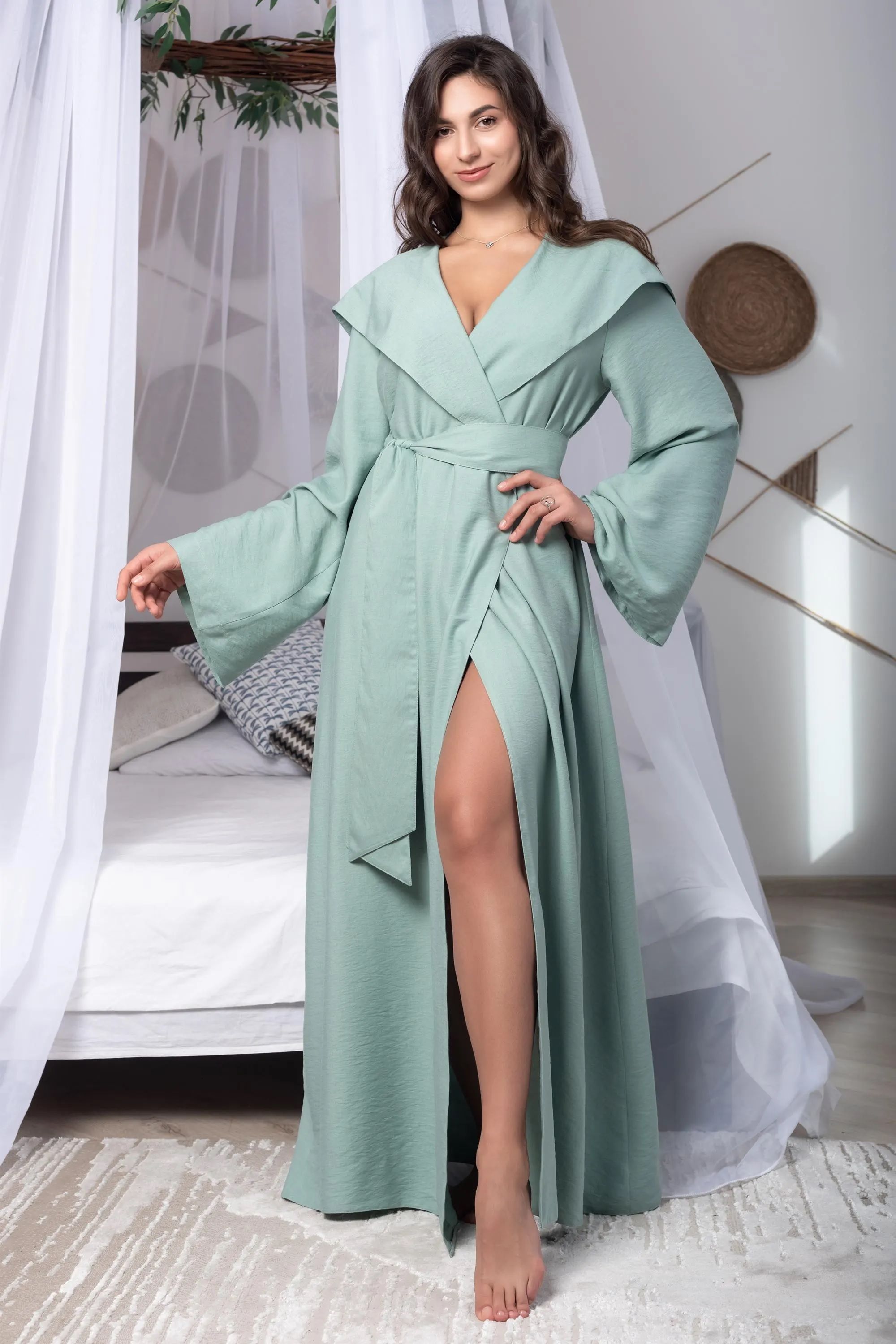 Woman wearing a light green dress with a slit in a room with white curtains and a bed.