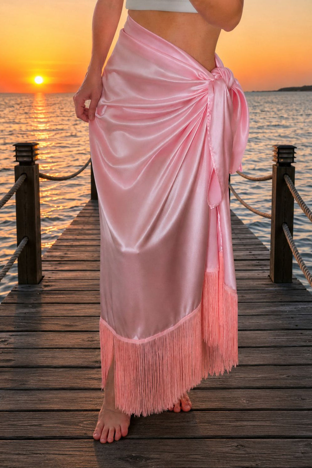 Person wearing a pink sarong with fringe on a wooden dock at sunset.
