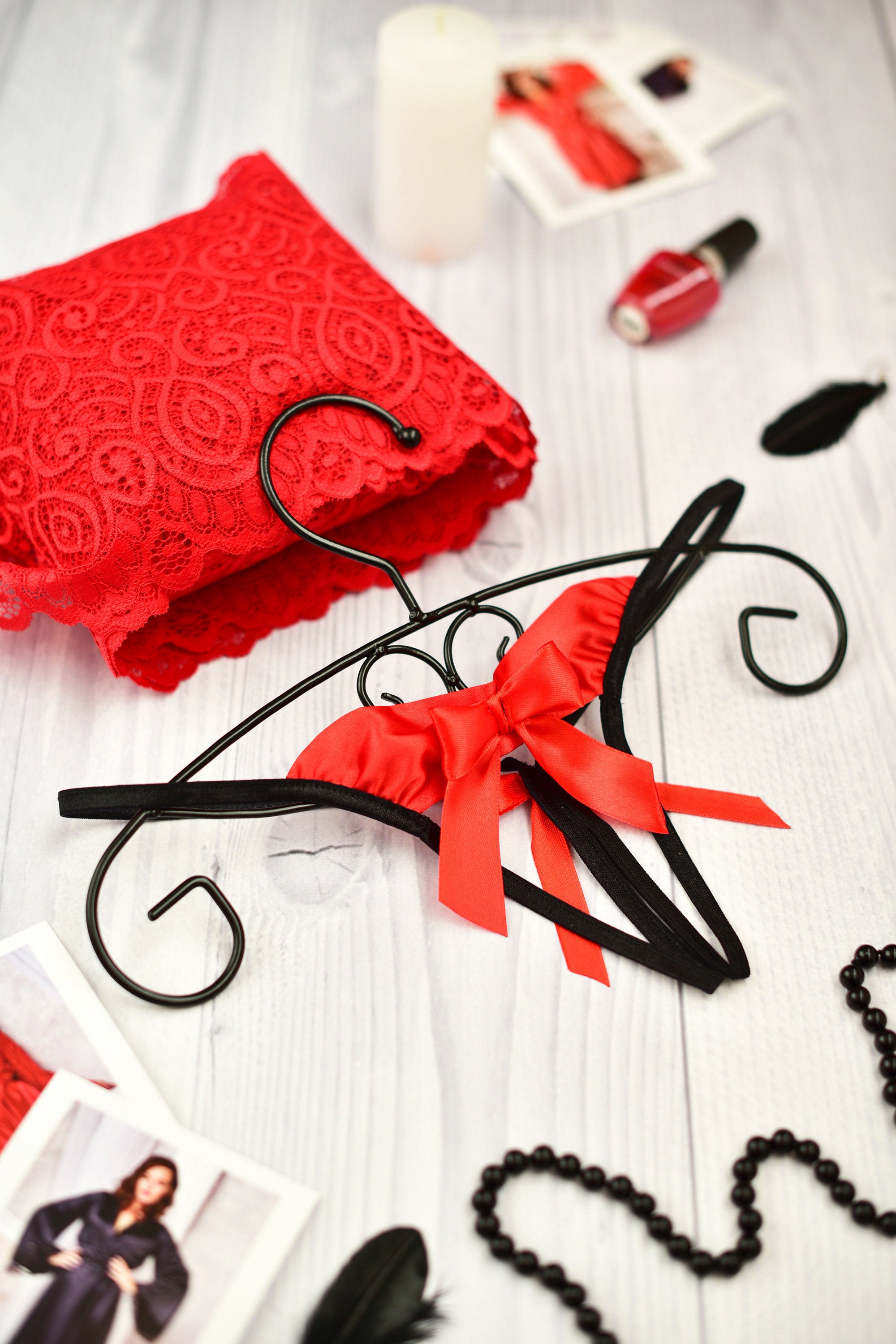 A black open string panty with a red bow, displayed on a wooden hanger, surrounded by red lace, nail polish, and other lingerie items.