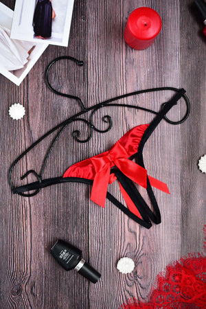 Red and black satin thong with a large red bow, displayed on a dark wood background with decorative props including a red candle, a black bottle, lace, and a photo frame.