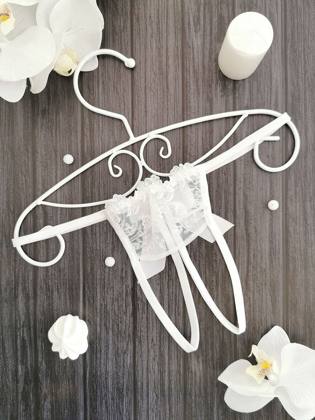 A white, stretch lace open crotch micro string thong panties with intricate detailing and a sheer design, displayed on a wooden surface surrounded by white flowers.