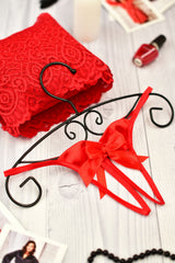 A sheer black open crotch string with satin bows, displayed on a white wooden surface with red lace, nail polish, and other accessories.