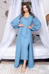 Woman wearing a long blue dress standing in a bedroom with white curtains and a bed.